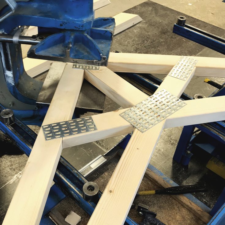 Pramoninis hidraulinis presas įkala metalinę jungimo plokštelę į medinės santvaros mazgą.